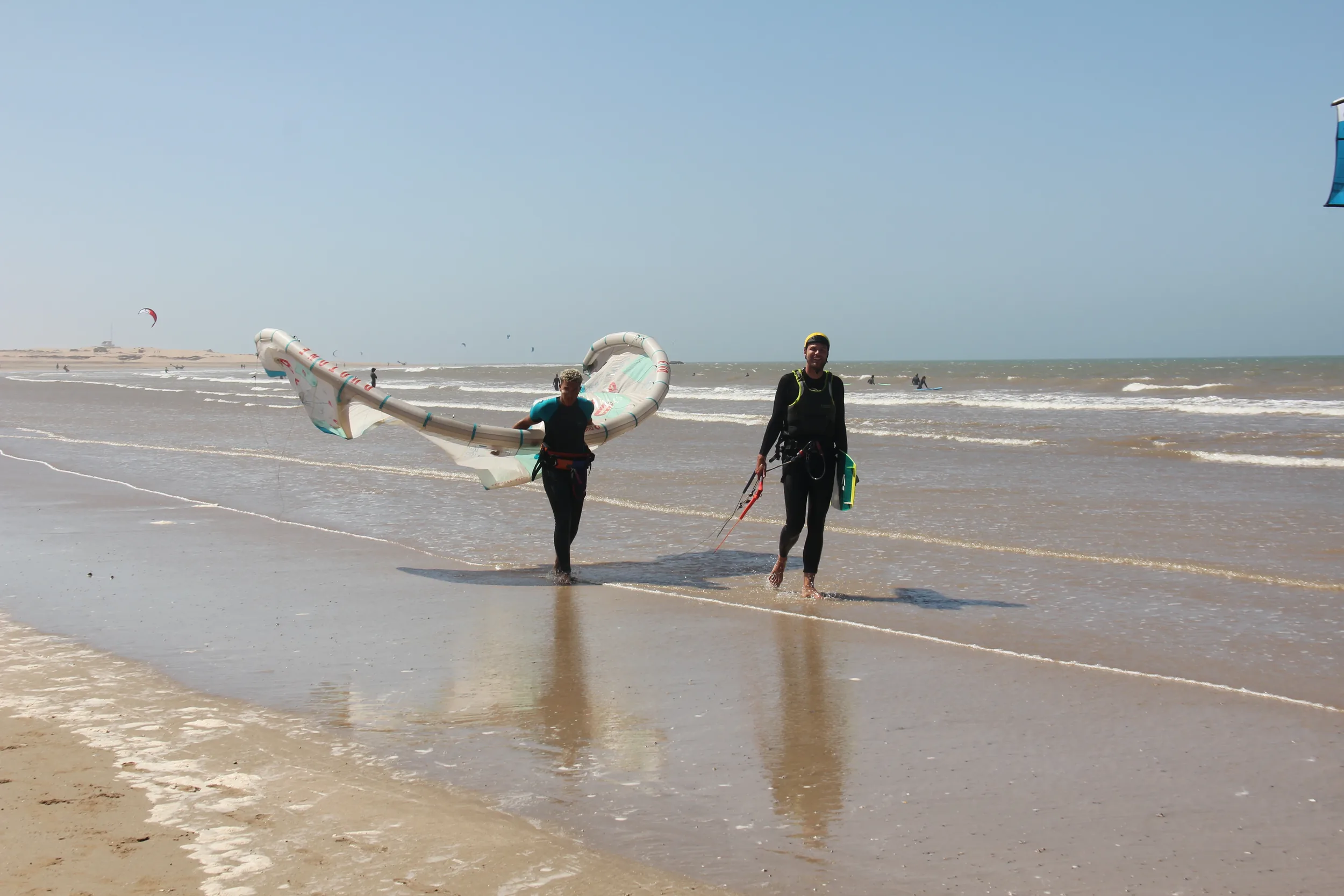 Deux kitesurfeurs marchant sur la plage humide avec leur aile, reflets sur le sable, Essaouira