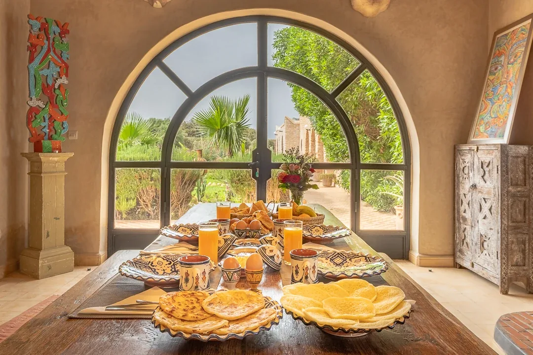 Table de petit-déjeuner dressée devant une fenêtre cintrée donnant sur le jardin, crêpes marocaines et jus d'orange