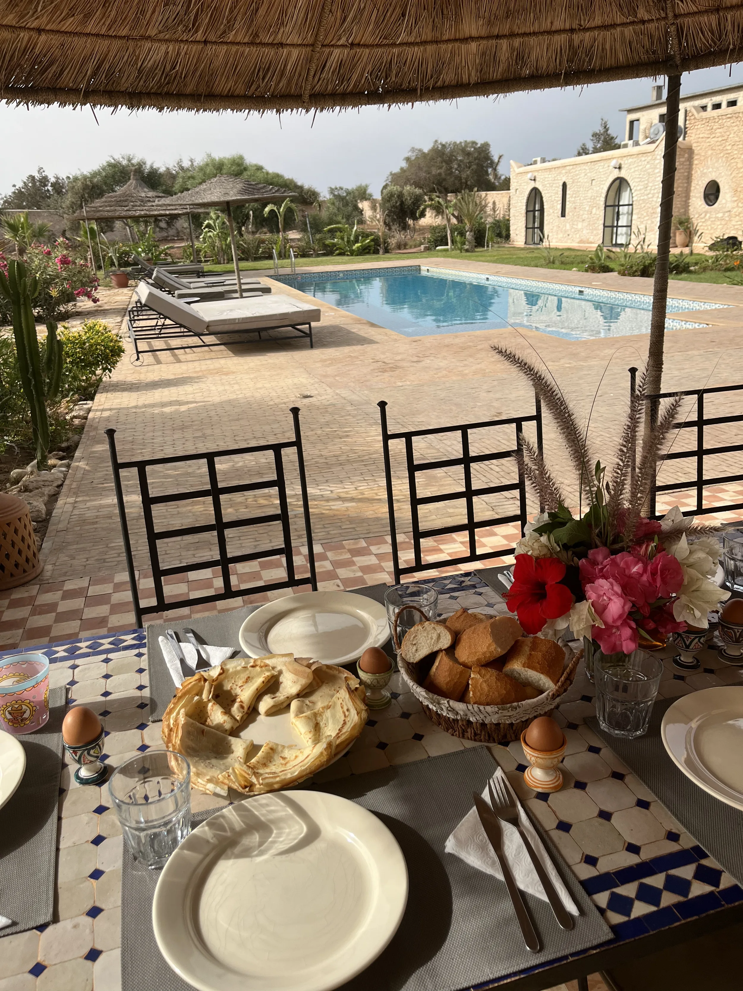 Table de petit-déjeuner dressée sous une paillote avec vue sur la piscine et le jardin