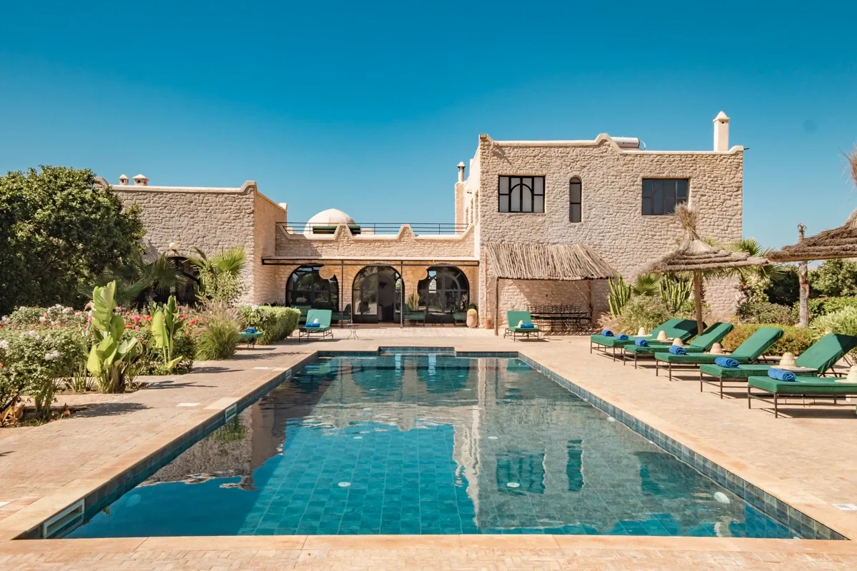 Longue piscine devant la façade en pierre de la villa avec terrasse couverte et transats