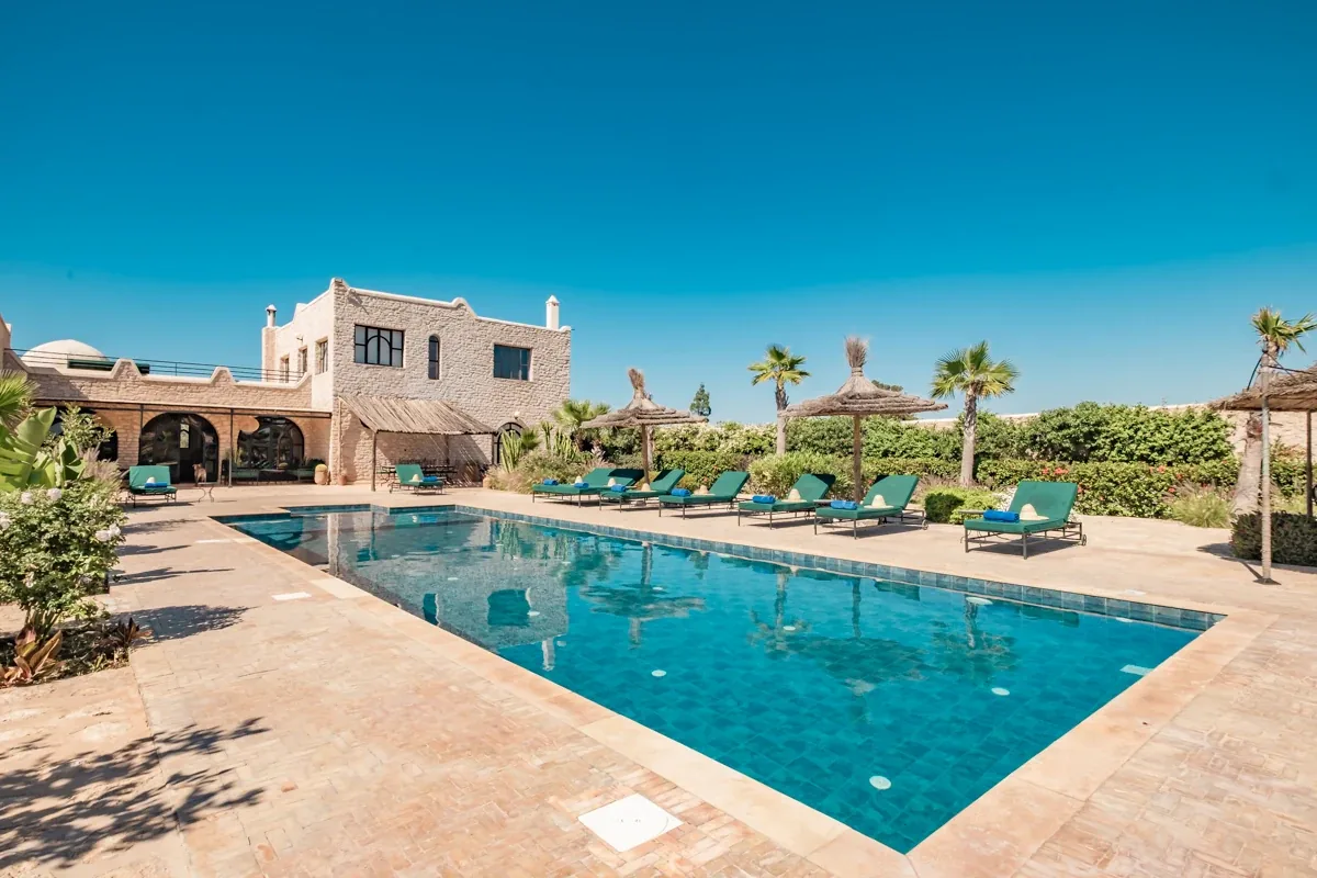 Piscine entourée de palmiers et de transats devant la villa à étage, grand jardin paysager