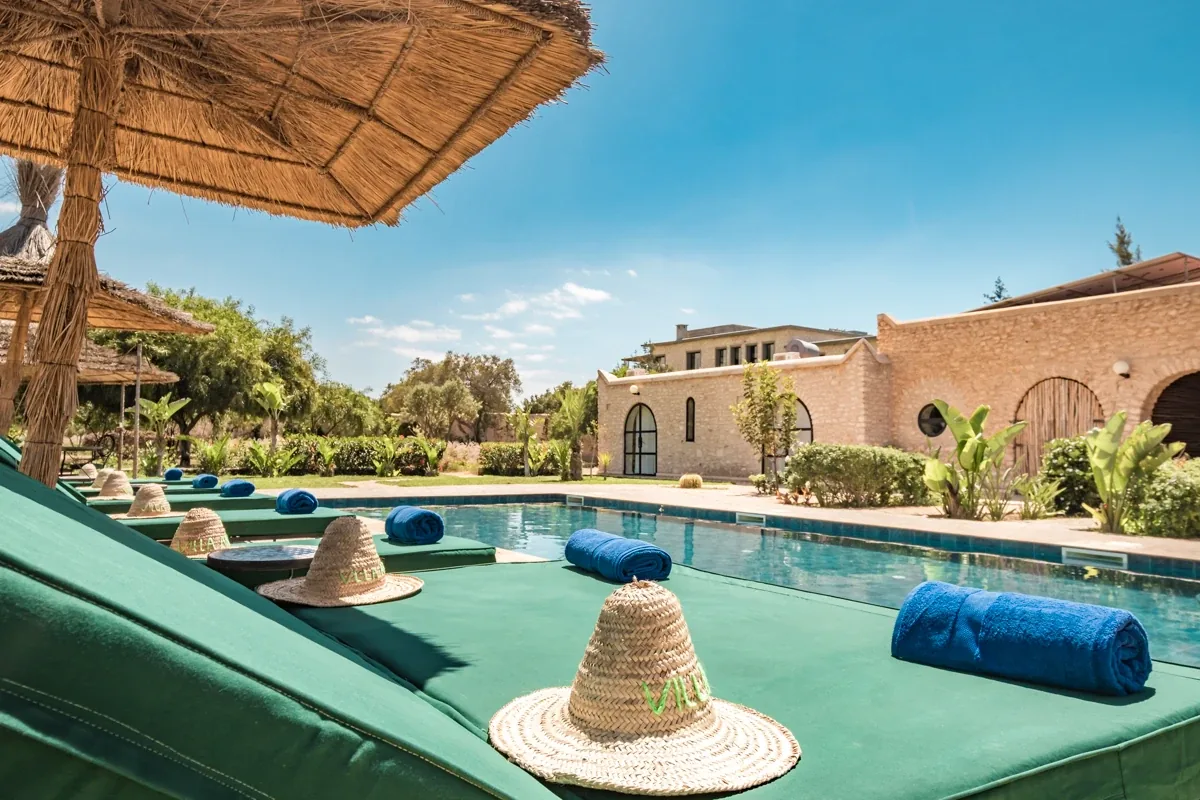 Espace de relaxation avec transats, coussins bleus et parasols en paille le long de la piscine