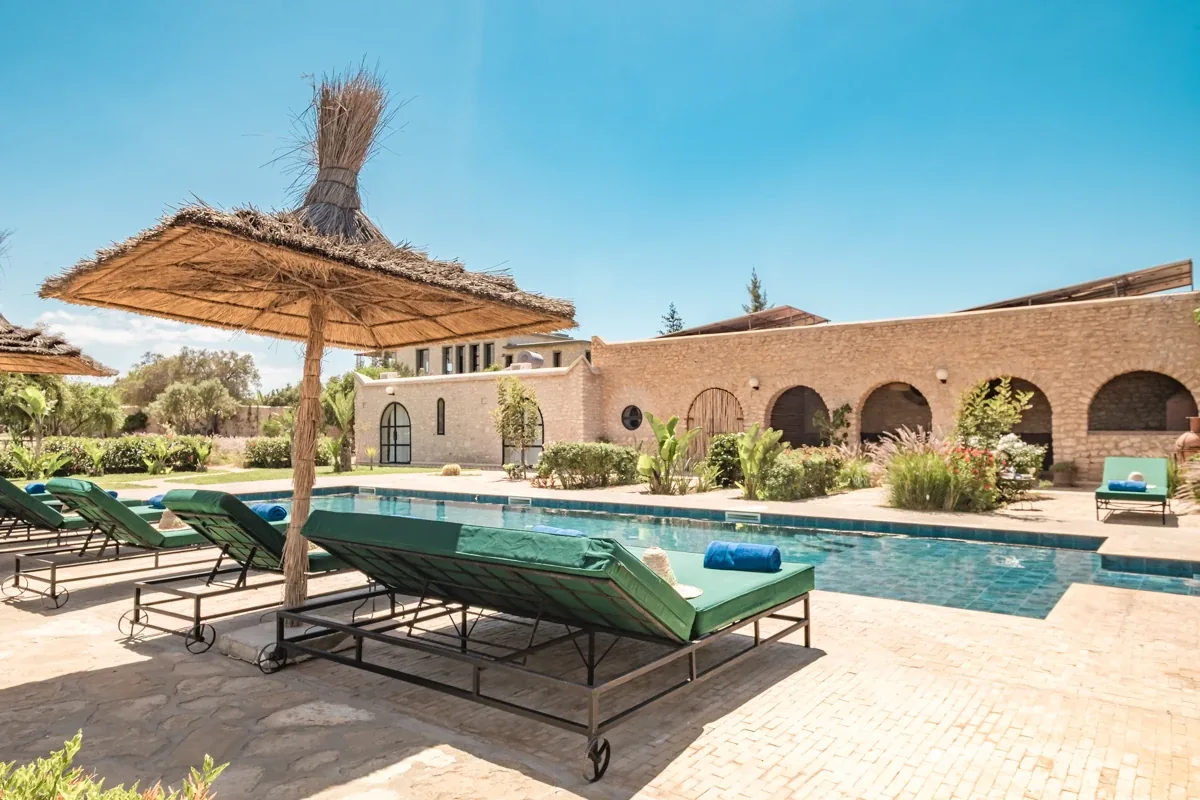 Transats au bord de la piscine sous un parasol en paille, terrasse ensoleillée de la villa
