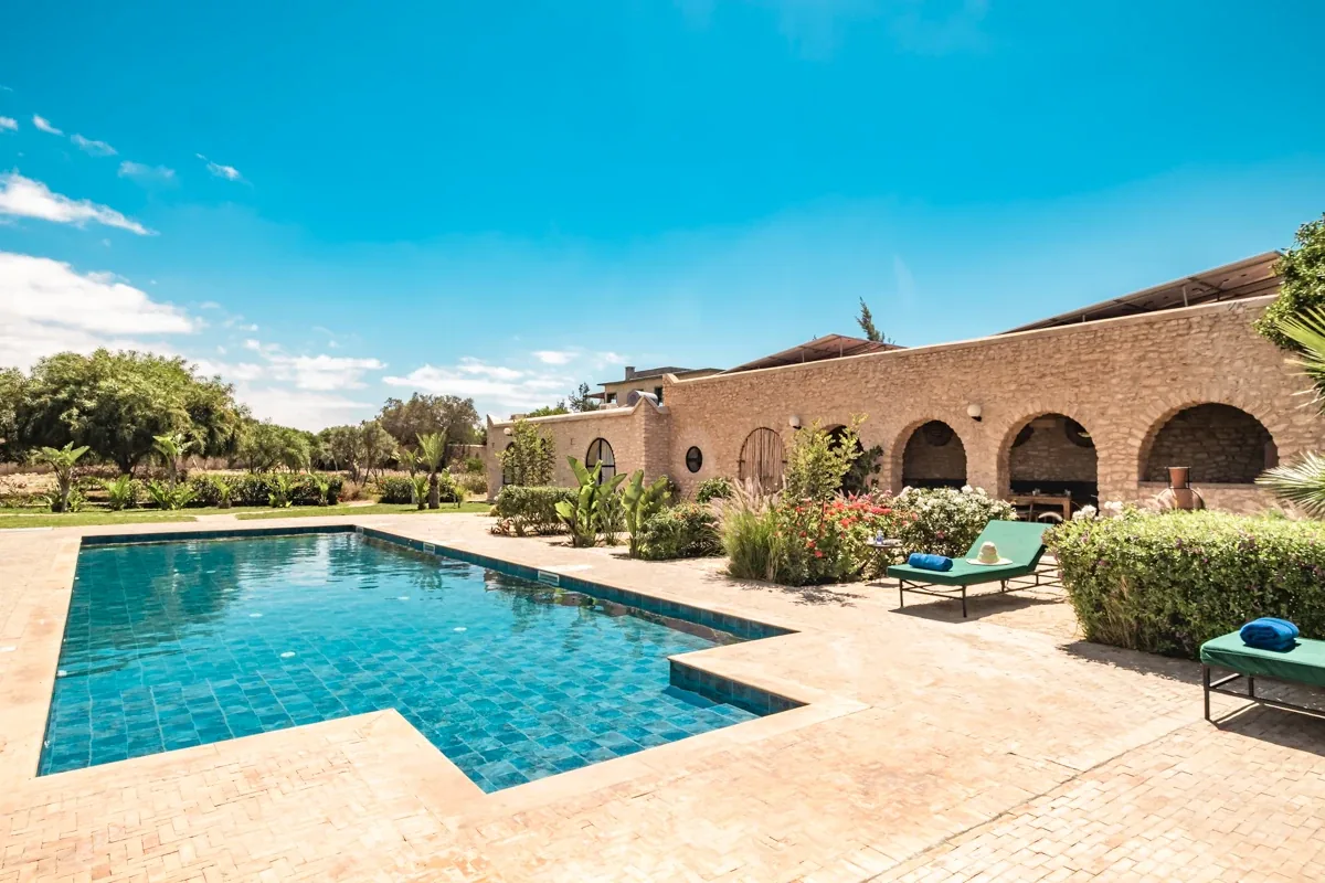 Grande piscine extérieure bordée de transats devant la villa aux arches en pierre, entourée d'un jardin verdoyant