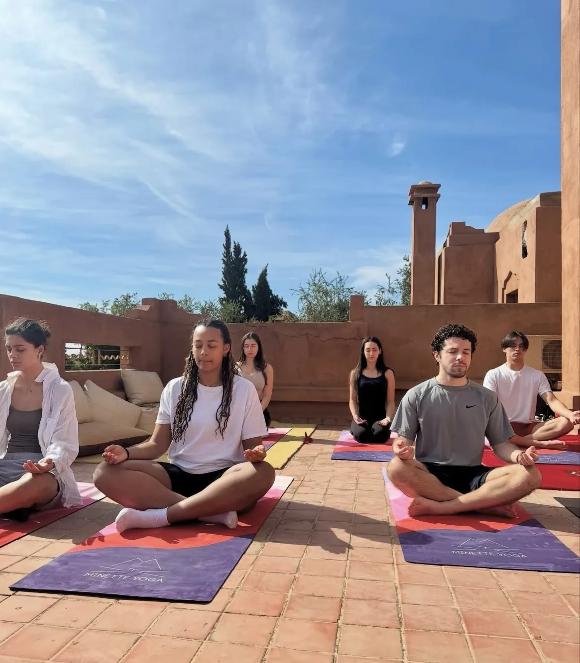 Groupe en méditation en lotus lors d'un cours de yoga sur une terrasse en pisé sous un ciel bleu