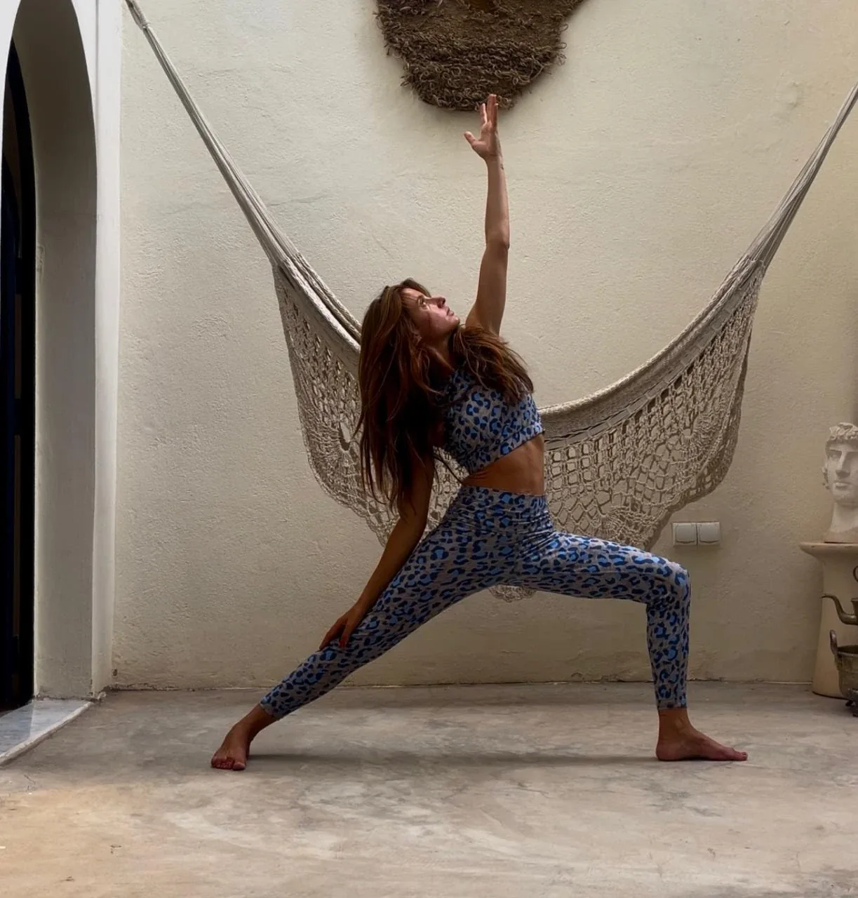 Femme en posture de yoga en fente devant un hamac dans un patio aux murs clairs