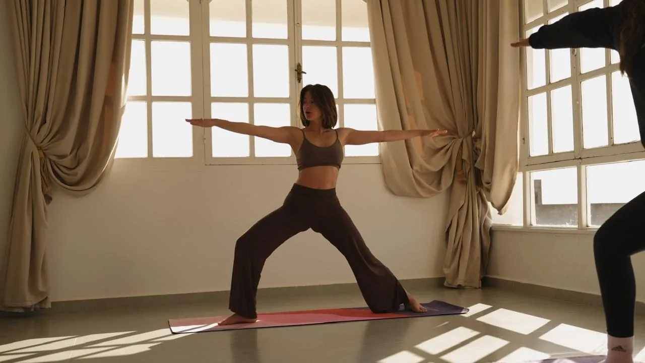 Femme en posture du guerrier sur un tapis de yoga dans une salle lumineuse aux grandes fenêtres